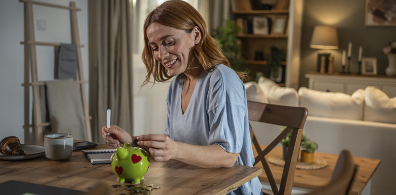 Woman looking at green piggy bank on table with open notebook next to her
